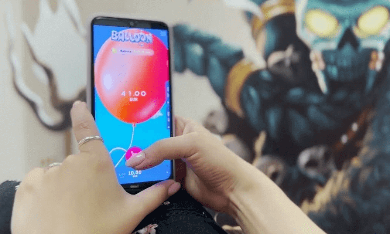 žena drží smartfón a hrá hru balloon crash s balónom s aktuálnou hodnotou .00$ eur na obrazovke.